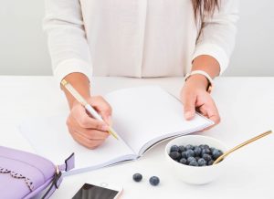 Lady writing in a notebook with blueberries on the table and a purple wallet